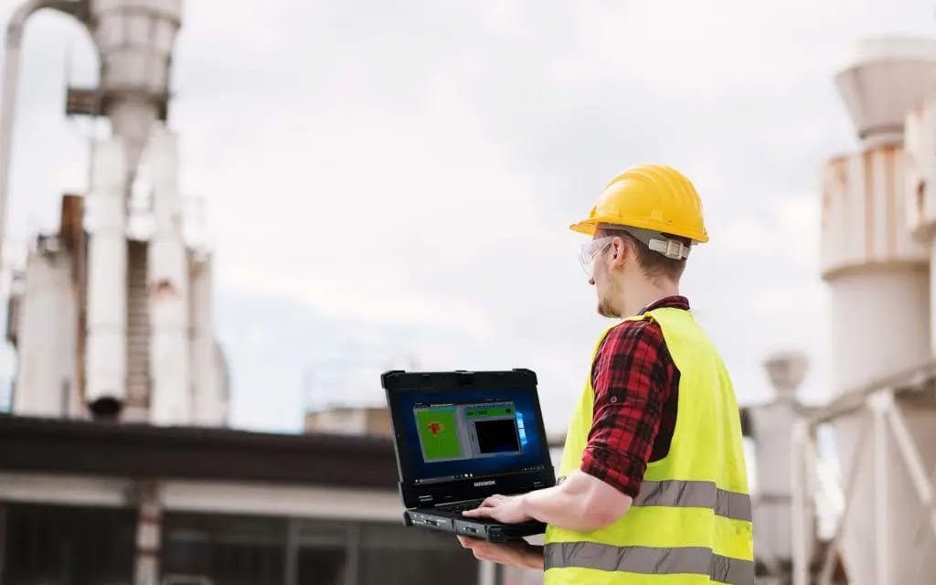 Technicien en gilet de sécurité utilisant un laptop durci sur un site industriel